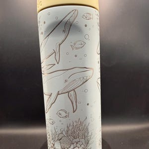 May include: A light blue water bottle with a white and yellow lid. The bottle features etched illustrations of whales, fish, bubbles, and coral. The design is a repeating pattern around the bottle.