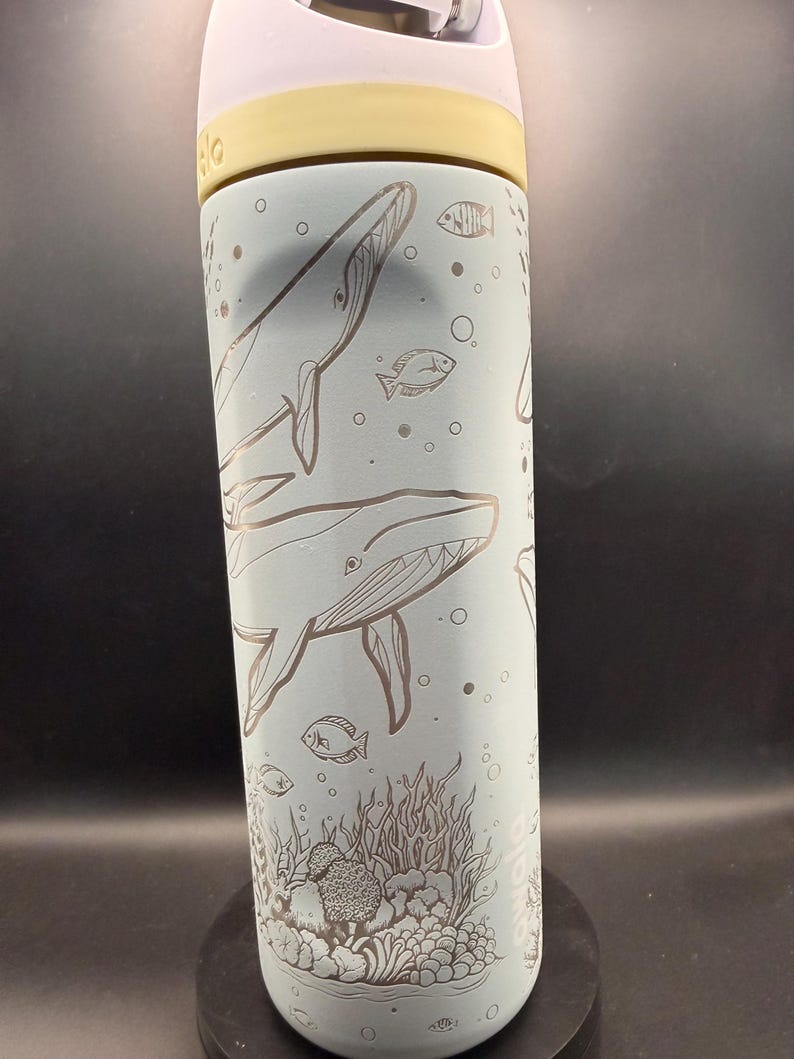 May include: A light blue water bottle with a white and yellow lid. The bottle features etched illustrations of whales, fish, bubbles, and coral. The design is a repeating pattern around the bottle.
