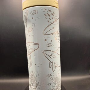 May include: A light blue water bottle with a white and yellow lid. The bottle features etched illustrations of whales, fish, and coral. The bottle is designed for cold beverages and has a handle for easy carrying.