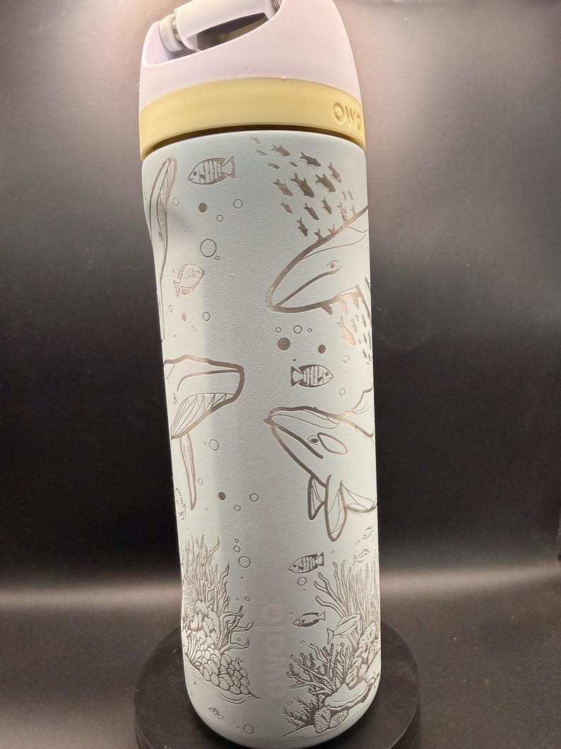 May include: A light blue water bottle with a white and yellow lid. The bottle features etched illustrations of whales, fish, and coral. The bottle is designed for cold beverages and has a handle for easy carrying.