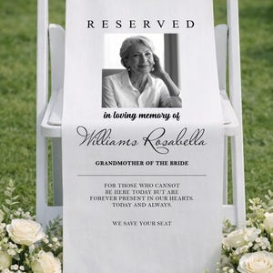 Pode incluir: Cadeira branca com uma faixa comemorativa que diz "RESERVED" e "in loving memory of Williams Rosabella, Grandmother of the Bride". A faixa inclui uma foto e uma mensagem. Arranjos florais brancos ladeiam a cadeira.