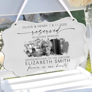 Può includere: Un cartello bianco con bordi smerlati con la scritta "Olivia & Henry 8.11.2025 riservato in memoria affettuosa" e una foto di una donna. Il cartello è appeso a una sedia bianca, con composizioni floreali sullo sfondo. Il cartello recita anche "Nonna dello sposo Elizabeth Smith per sempre nei nostri cuori."
