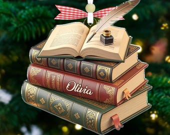 May include: A festive Christmas tree ornament depicting a stack of books. The top book is open, with a quill and inkwell. The books are in shades of red, green, and brown, with gold detailing. The name "Olivia" is visible. A red and white ribbon is at the top.