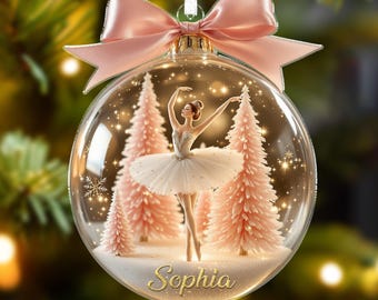 May include: A clear glass Christmas ornament featuring a ballerina in a white tutu and pink trees. The ornament has a pink bow and the name "Sophia" in gold script. The background is a blurred Christmas tree.