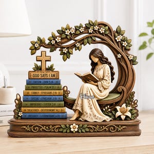 Può includere: Una statuetta decorativa che raffigura una persona seduta che legge un libro sotto un ramo d'albero curvo con fiori bianchi. Accanto si trova una pila di sette libri con titoli come God Says I Am, Chosen, Protected, Family, Special, Important, Treasured e Precious, sormontata da una croce di legno.