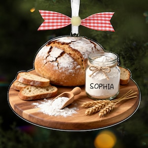 Op de afbeelding: Een ronde houten ornament met een brood, sneetjes brood, bloem en een pot met de naam "SOPHIA". Een houten lepel en tarwearen zijn ook aanwezig. Het ornament is versierd met een rood-wit geruite strik.