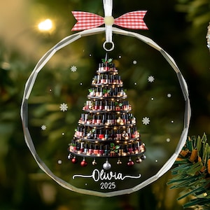 May include: A clear, round Christmas ornament with a nail polish Christmas tree design. The tree is made of various nail polish bottles, decorated with small ornaments and lights. The ornament has a red and white bow and the name "Olivia" and the year "2025" written on it.
