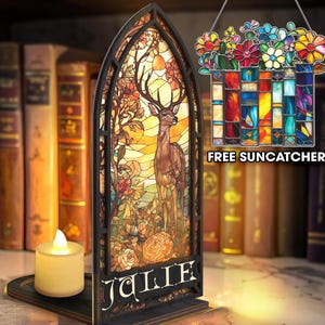 Op de afbeelding: Een decoratief verlicht stuk in de stijl van een glas-in-loodraam met een hert in een boslandschap. Het zwart omlijste stuk heeft de naam "JULIE" aan de basis. Een kleine kaars staat op een houten voet. Een hangende glas-in-lood bloemenvanger is ook inbegrepen.