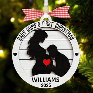 May include: A round, wooden Christmas ornament with the text "BABY BUMP'S FIRST CHRISTMAS" and "WILLIAMS 2025". The ornament features a black silhouette of a pregnant person and a person touching the baby bump, with a red heart. It is decorated with a red and white checkered bow.