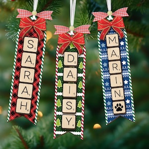 May include: Three personalised Christmas tree decorations with names spelled out in Scrabble tiles. Each decoration has a red bow and a ribbon for hanging. The decorations feature different patterns: plaid, green with trees, and blue with stars.