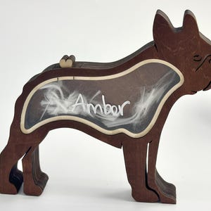 Może przedstawiać: Drewniane pudełko pamiątkowe w kształcie psa w odcieniach brązu, z napisem "Amber" w białym skrypcie. Pudełko ma przezroczyste okno wypełnione białym puchem i mały detal w kształcie serca na górze.