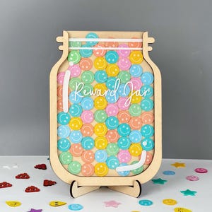 May include: A wooden reward jar with a clear front and a stand. The jar is filled with colorful smiley face stickers. The text "Reward Jar" is written on the front of the jar.