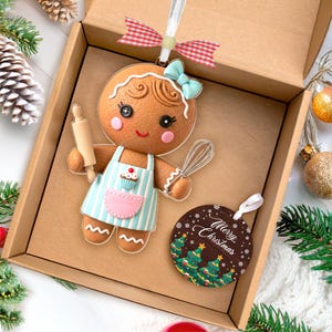 May include: A gingerbread woman Christmas ornament with a light blue bow and apron, holding a rolling pin and whisk. A round ornament with "Merry Christmas" text and Christmas trees is also included.