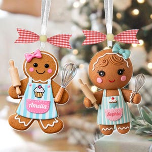 May include: Two gingerbread cookie ornaments with white ribbon hangers and red and white checkered bows. Each ornament has a pink bow, a blue and white striped apron, and holds a rolling pin and whisk. One apron says "Amelia", the other says "Sophia".