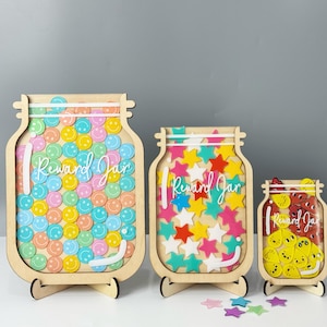 May include: Three wooden reward jars with stands. The largest jar is filled with colorful smiley face stickers, the middle jar is filled with colorful star stickers, and the smallest jar is filled with colorful heart stickers. Each jar has the words "Reward Jar" written on it.