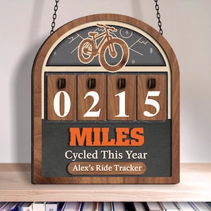 May include: Wooden bicycle ride tracker displaying "0215 Miles Cycled This Year" and "Alex's Ride Tracker". The top has a bicycle graphic and mountain scene. The sign is suspended by chains.