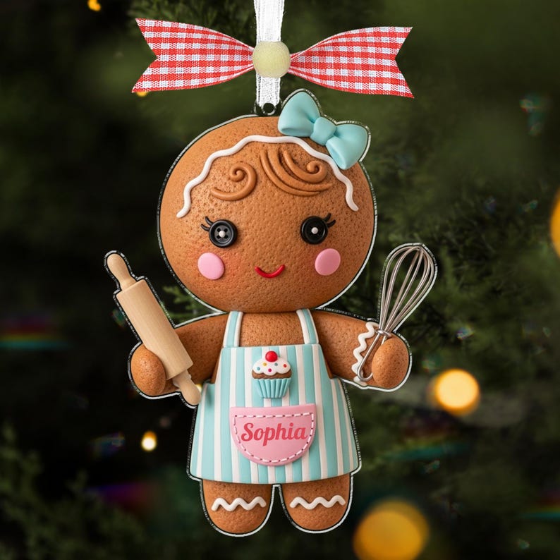 May include: A gingerbread woman Christmas ornament with a light blue bow and apron. The apron has blue stripes and a cupcake design. The ornament is holding a rolling pin and a whisk. The name "Sophia" is on the apron.