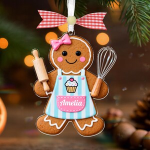 May include: Gingerbread cookie ornament with a pink bow, holding a rolling pin and whisk. The ornament has a blue and white striped apron with a cupcake design and the name "Amelia". A red and white checkered bow is at the top.