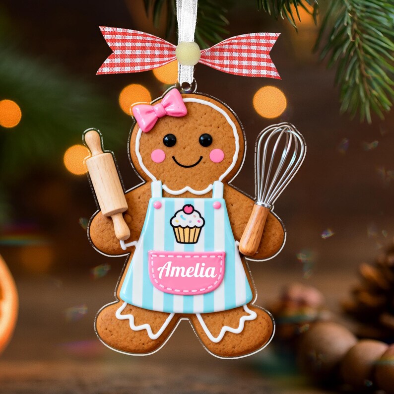 May include: Gingerbread cookie ornament with a pink bow, holding a rolling pin and whisk. The ornament has a blue and white striped apron with a cupcake design and the name "Amelia". A red and white checkered bow is at the top.