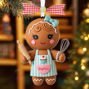 May include: A gingerbread-person Christmas ornament with a blue bow, apron, and rolling pin. The apron has blue stripes and a cupcake design. The ornament is personalized with the name "Sophia" and is decorated with a red and white checkered bow.