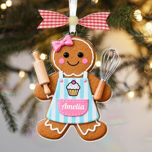 May include: Gingerbread cookie ornament shaped like a baker, wearing a blue apron with a cupcake design and the name "Amelia". The ornament has a pink bow, rolling pin, and whisk. It hangs from a red and white ribbon.