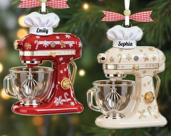 May include: Two Christmas ornaments shaped like stand mixers, one red and one cream-colored. Each has a chef's hat with a name, a clear bowl, and a whisk. The red mixer has white snowflake and candy cane details. The cream mixer has gold accents.