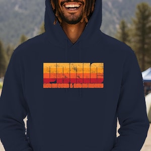 May include: Navy blue hoodie with a retro-style graphic that reads "Idaho" in orange and yellow, with the text "Ride, Camp & Get Money" below. The hoodie has a drawstring hood and a front pocket. The background shows a mountain range and camping scene.