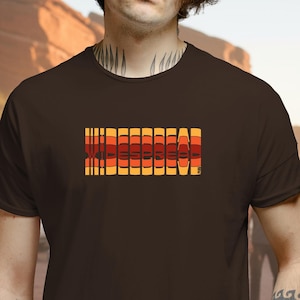 May include: A brown t-shirt featuring the word "WIDESPREAD" in a retro, block-letter style. The text uses a gradient of orange, yellow, and red hues. The shirt is a solid brown color.
