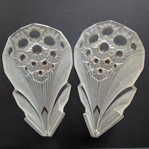 May include: Two Art Deco style glass panels with a floral design. Each panel is clear with etched details, featuring a fan-shaped base and a top section with circular cutouts. The panels are symmetrical and appear to be decorative.