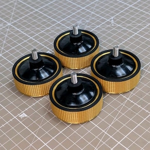 Puede incluir: Cuatro patas de muebles negras y doradas con una base dorada texturizada, una parte superior negra y una varilla roscada plateada que se extiende hacia arriba. Las patas están dispuestas sobre una superficie marrón claro con líneas de cuadrícula.
