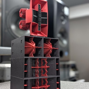 May include: A black and red speaker system with a distinctive design. The system comprises stacked black rectangular boxes with red internal components, topped by a large red horn-shaped speaker. The design is modern and geometric.