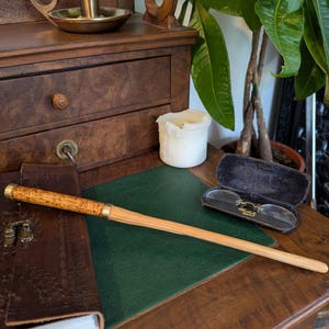 Op de afbeelding: Een lichtbruine houten toverstaf met messing details ligt op een groen bureau. Een leren notitieboek en een bril in een etui zijn ook aanwezig. Een kaars en een messing kandelaar staan op een houten bureau met laden.