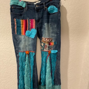 May include: Dark wash denim jeans with colorful patches and turquoise accents. The jeans feature a heart-shaped patch, fringed bell bottoms, and turquoise lace and fringe details. The jeans are hanging on a hanger.