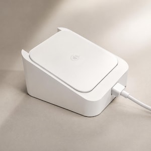May include: A white, angled charging dock with a square top surface featuring a small circular logo in the center. A white charging cable is plugged into the side of the device, extending outward from the base of the unit on a neutral surface.