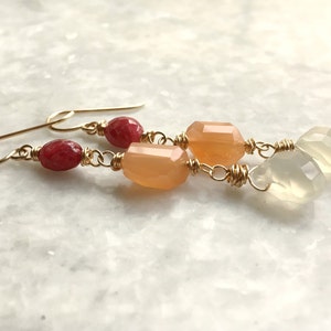 Puede incluir: Pendientes colgantes dorados con piedras preciosas rojas, melocotón y blancas. Los pendientes tienen un diseño delicado con cuentas facetadas y redondeadas, conectadas por alambre dorado.