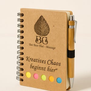 Könnte beinhalten: Ein Spiralnotizblock mit einem Kraftpapier-Einband und einem Bambusstift. Der Einband zeigt ein Logo und den Text "Kreatives Chaos beginnt hier." Das Notizbuch enthält Haftnotizen in Gelb, Rosa, Grün, Orange und Blau.