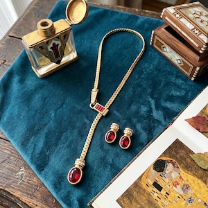 May include: A gold-tone lariat necklace featuring a curb chain, a rectangular bar with three red baguette stones, and a dangling oval red pendant. The set includes matching oval red stud earrings with small clear crystals at the top.