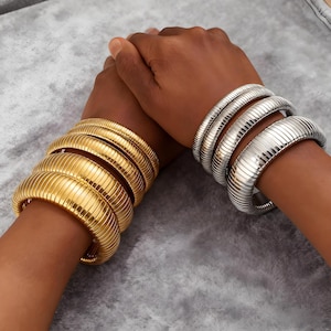 May include: Gold and silver-toned, ribbed metal bracelets. The gold bracelets are on the left, and the silver bracelets are on the right. The bracelets are thick and have a rounded, tubular design. The bracelets are worn on the wrists.