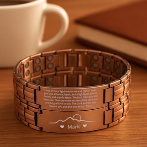 May include: A copper-colored bracelet with a rectangular link design. The bracelet has an engraved message and a mountain and heart design with the name "Mark". A white coffee mug and a brown book are in the background.