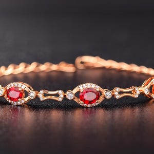 Può includere: Un bracciale in oro rosa con pietre preziose rosse ovali e piccole pietre bianche. Il bracciale ha una fascia intrecciata. Le pietre rosse sono circondate da pietre bianche più piccole, su sfondo scuro.