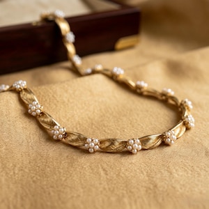 Puede incluir: Un collar de metal dorado que presenta una serie de eslabones texturizados en forma de hoja que se alternan con pequeños grupos de perlas blancas redondas de imitación. El collar tiene un diseño articulado y flexible en curva.