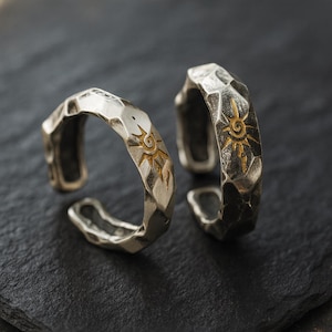 May include: Two silver-colored adjustable rings with a hammered texture. Each ring features a sun design in gold. The rings are displayed on a dark, textured surface, showcasing their intricate details and craftsmanship.