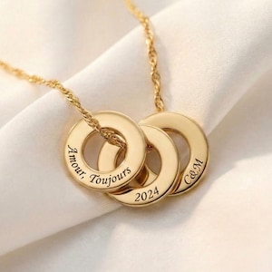 Könnte beinhalten: Eine goldfarbene Halskette mit drei kreisförmigen Anhängern. Jeder Anhänger hat ein Loch in der Mitte und ist mit Text graviert. Der erste Anhänger trägt die Aufschrift "Amour, Toujours", der zweite "2024" und der dritte "C&M".