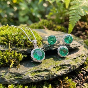 May include: A jewelry set featuring a pendant necklace, a pair of earrings, and a ring. Each piece has a large, round, emerald-green gemstone surrounded by small, clear stones. The jewelry is set on a stone surface with moss.