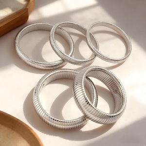 May include: Six silver-coloured, flexible coil bracelets are displayed. The bracelets are circular with a ribbed texture, reflecting light. The jewellery is presented on a white surface, with a wooden tray in the background.
