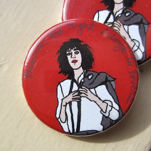 May include: Two red round buttons with a graphic of a person in a white shirt and black jacket. The buttons have the text "Because the night belongs to lovers" in a cursive font around the edge of the button.