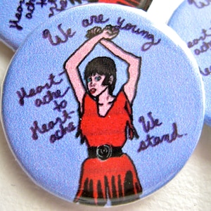 May include: A blue button with a woman in a red dress with black trim. The text on the button reads "We are young, Heart ache to Heart ache, We stand."