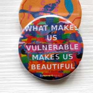Pode incluir: Um botão colorido com o texto "What makes us vulnerable makes us beautiful" e o nome "Brando Brown" na parte inferior.
