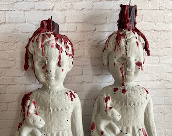May include: Two concrete statues of children, each holding a small animal. The statues are painted white with red dripping accents, and have black candles on top. The statues are placed in front of a white brick wall.