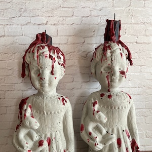May include: Two concrete statues of children, each holding a small animal. The statues are painted white with red dripping accents, and have black candles on top. The statues are placed in front of a white brick wall.